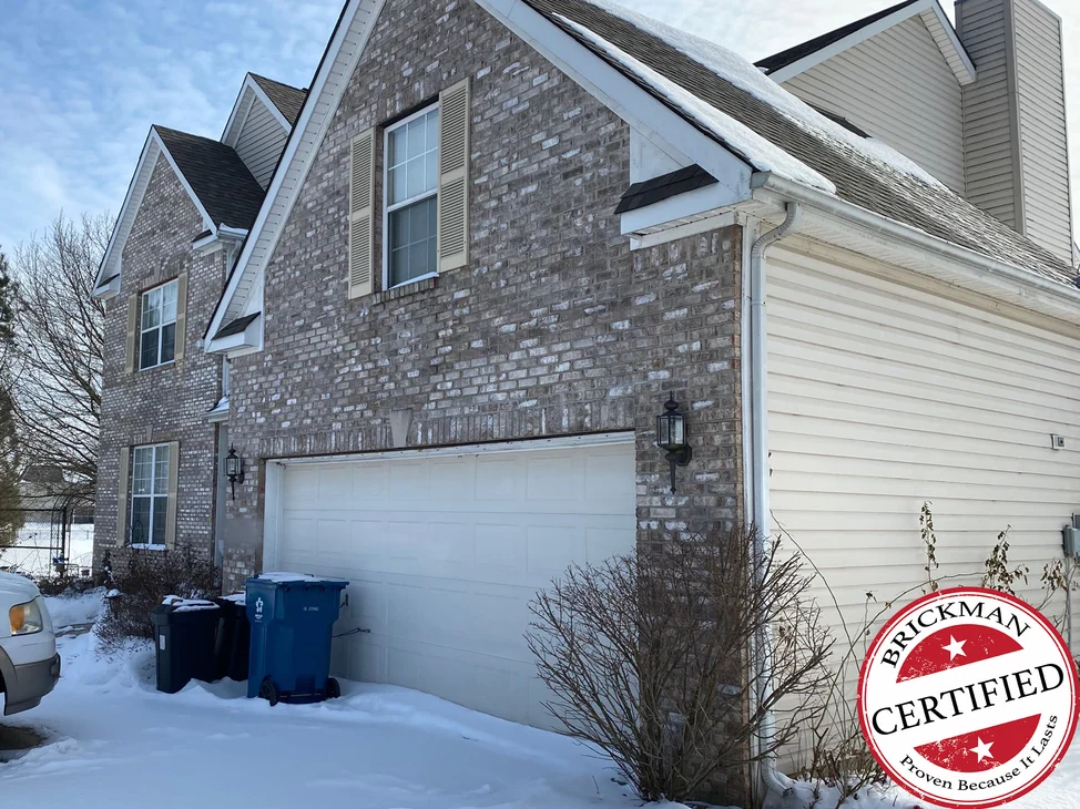 The garage wing and the front gable of the house in 2021 under snow, the brick one grey and cream range from the upper window down to the pier beside the garage door, with the certification stamp in the corner