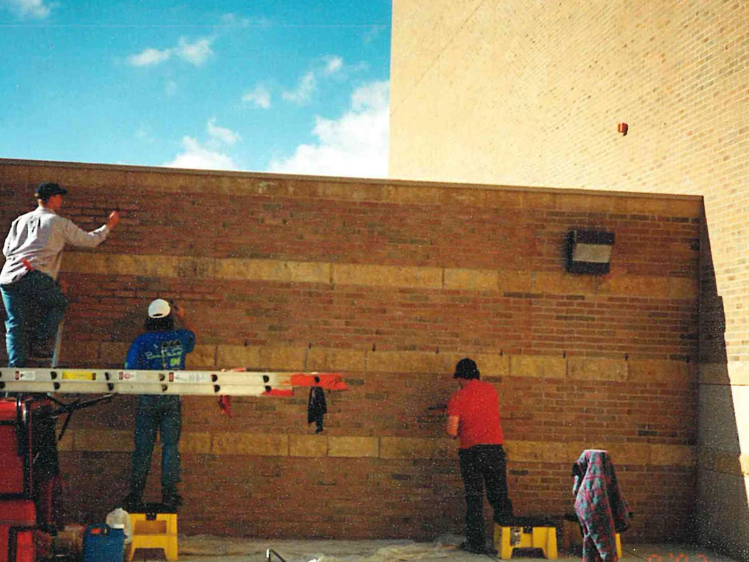 Three applicators working across a long banded wall from a ladder and step stools, red brick between wide courses of lighter brick, a taller tan brick wall behind