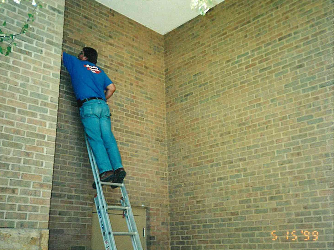 An applicator on an extension ladder working the top of a tall buff brick return, the frame carrying a burned-in camera date reading 5 15 99