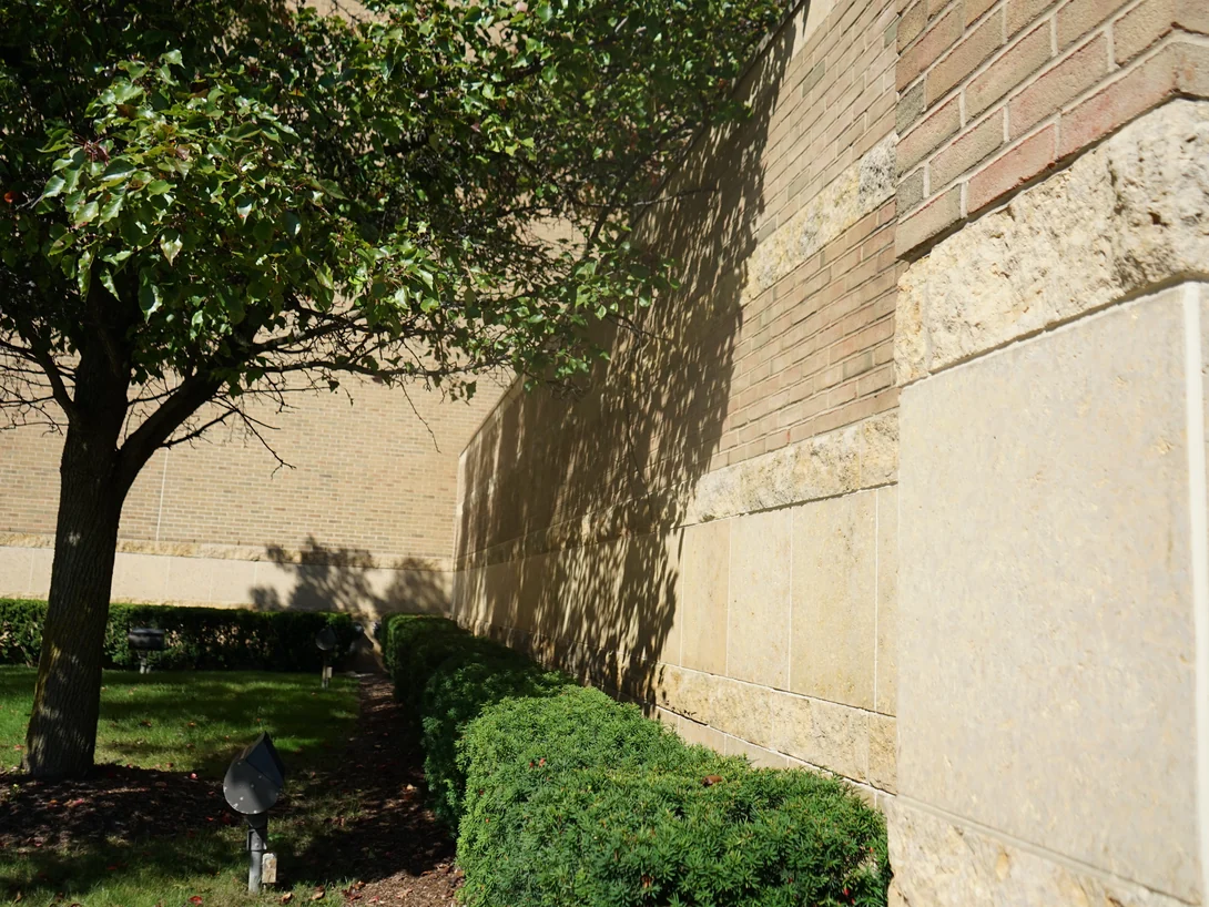 A long tan brick elevation of the mall in 2019 running away under a tree to an outside corner faced in limestone blocks, red and tan brick above the stone base
