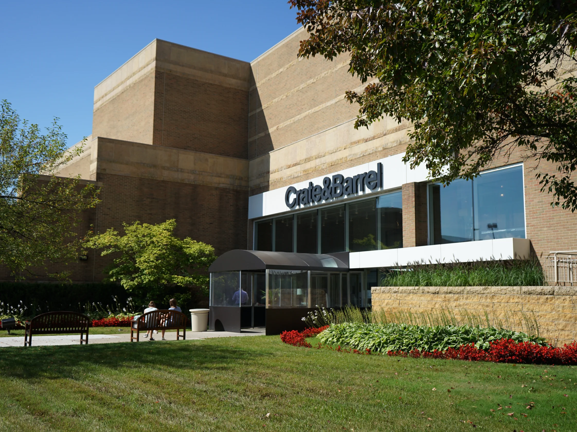 The mall in 2019 seen from the lawn, tan brick stepping up in blocks behind a Crate & Barrel storefront and a glazed entrance vestibule