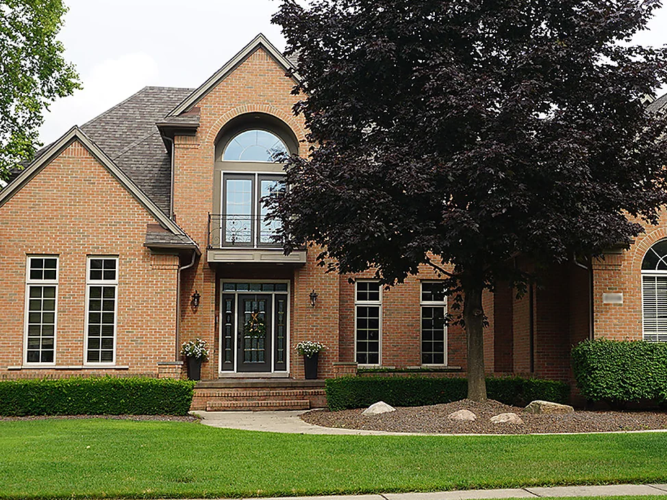 The front of the two-story house in Utica in 2019, one even range of salmon and tan brick around an arched window over the entrance, a dark leafed maple beside it