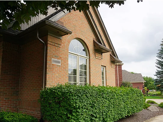 A gable end of the house in 2019, salmon and tan brick around an arched window over a clipped hedge, with a spruce and the street beyond