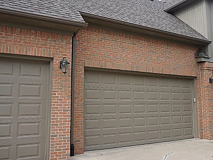 The attached garage in 2019, two tan doors set in salmon and tan brick under a grey sided upper story, the brick reading as one range across the piers