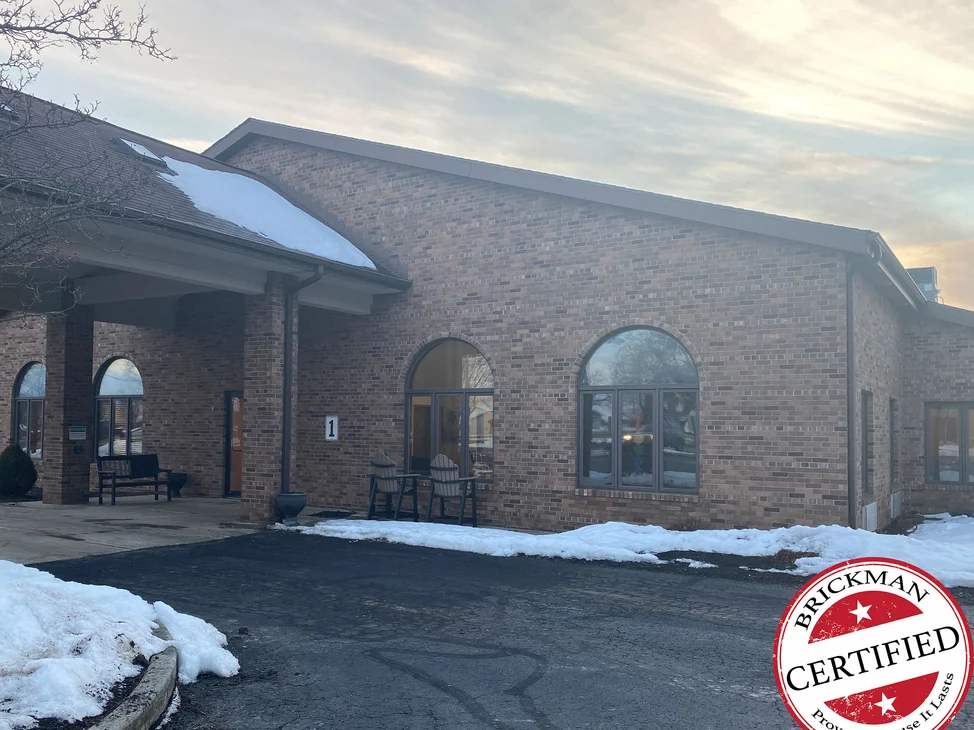 The building in 2021 with snow on the ground, a new covered entrance built across the gable wall beside two arched windows, with the certification stamp in the corner