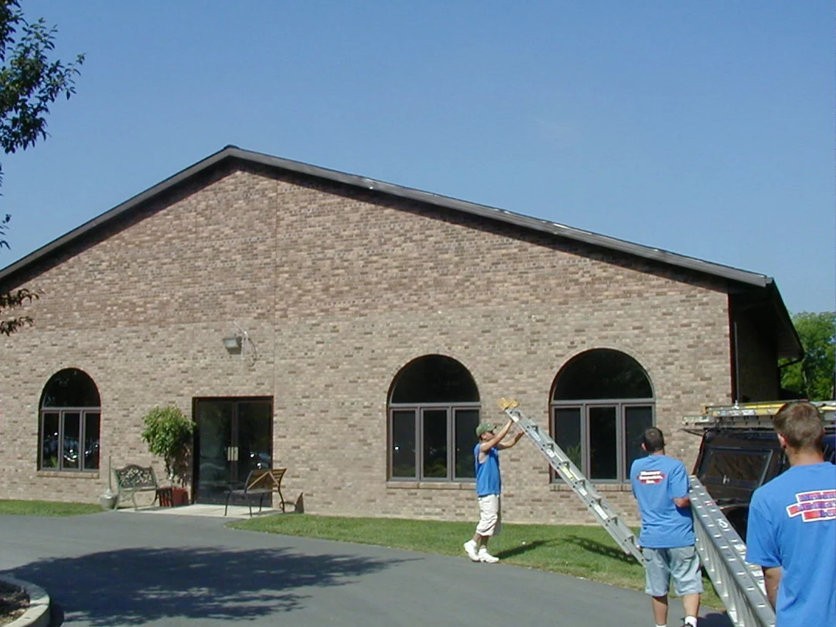 The gable end of the single-story building in 2004, the peak above the arched windows and entrance doors laid in a lighter run of brick than the wall below it, with three workers carrying an extension ladder across the drive
