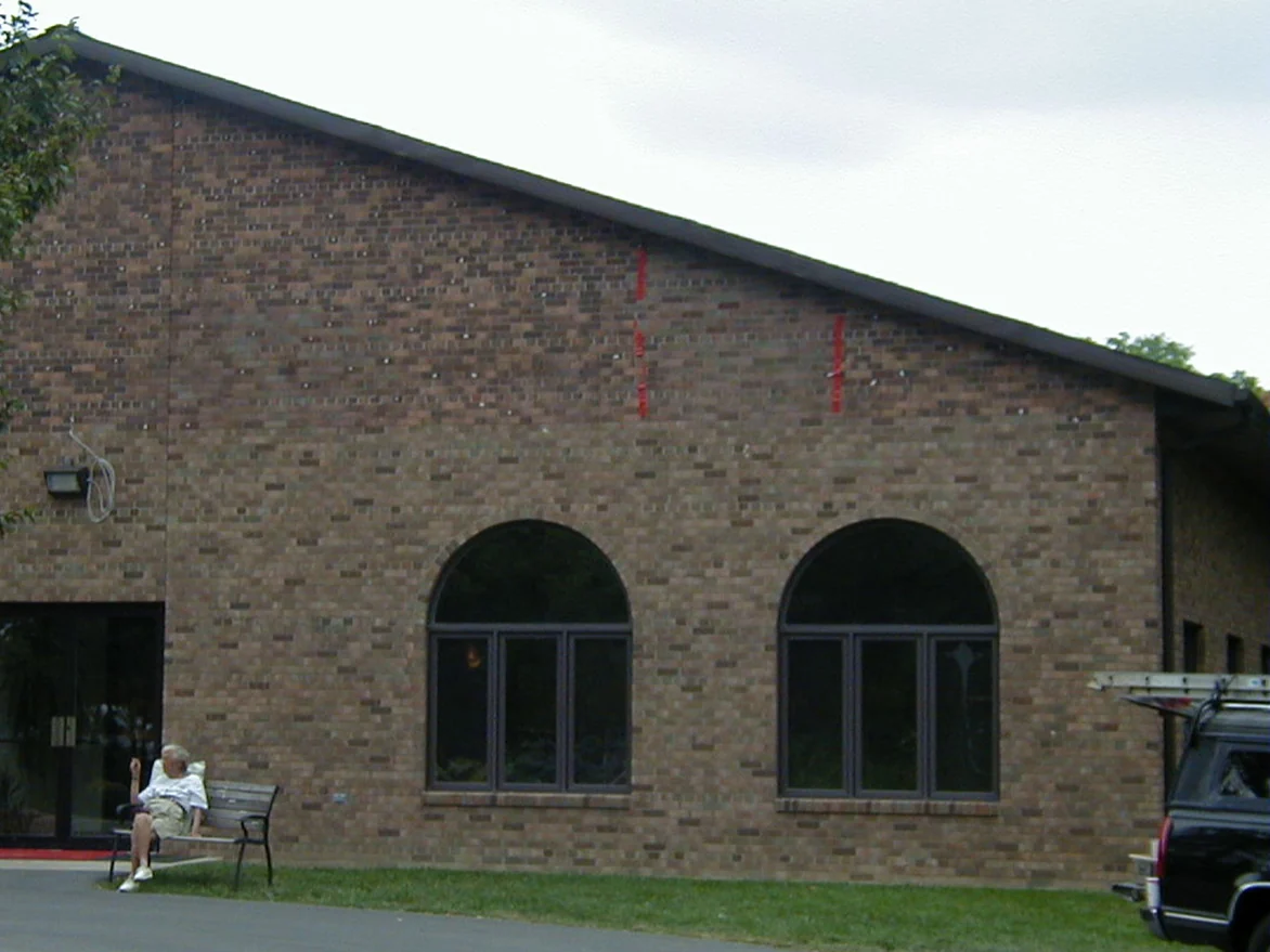 The same gable in 2004 with the peak brought darker to sit with the wall below, two strips of red tape marking courses high on the wall and a man on a bench by the entrance