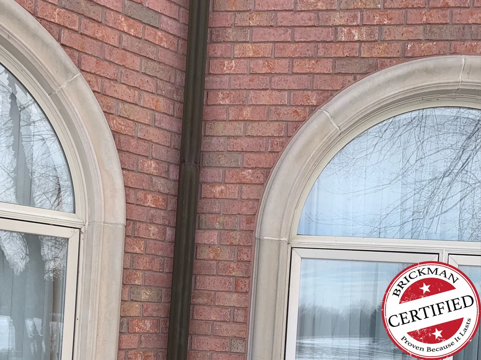 The inside corner in 2021 at a dark downspout between two arched windows, red brick running one range around the turn, with the certification stamp in the lower right corner