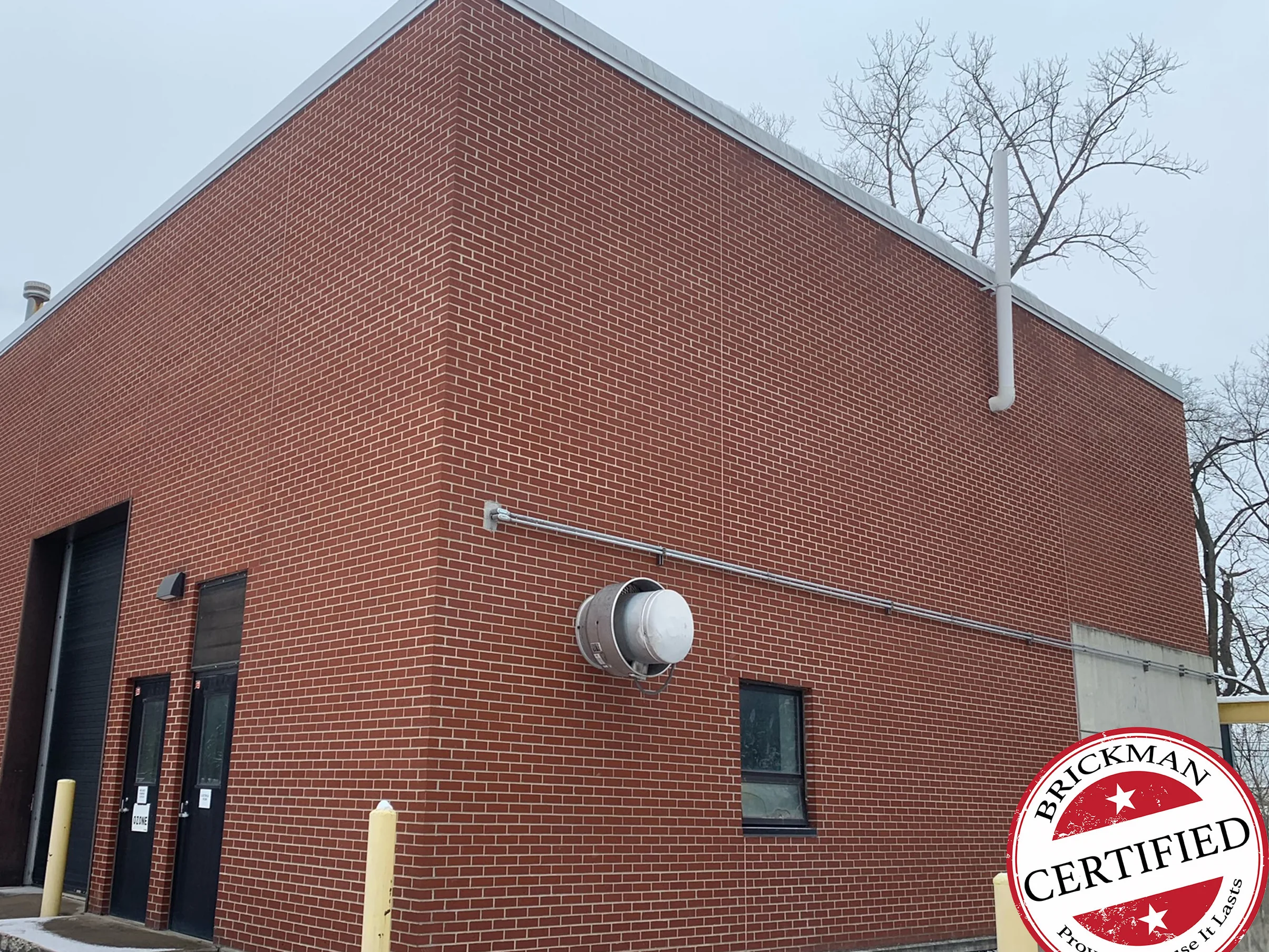 The same corner in 2021 with snow on the ground, one range of red brick across both walls, a round exhaust vent and a window on the near wall, and the certification stamp in the lower corner