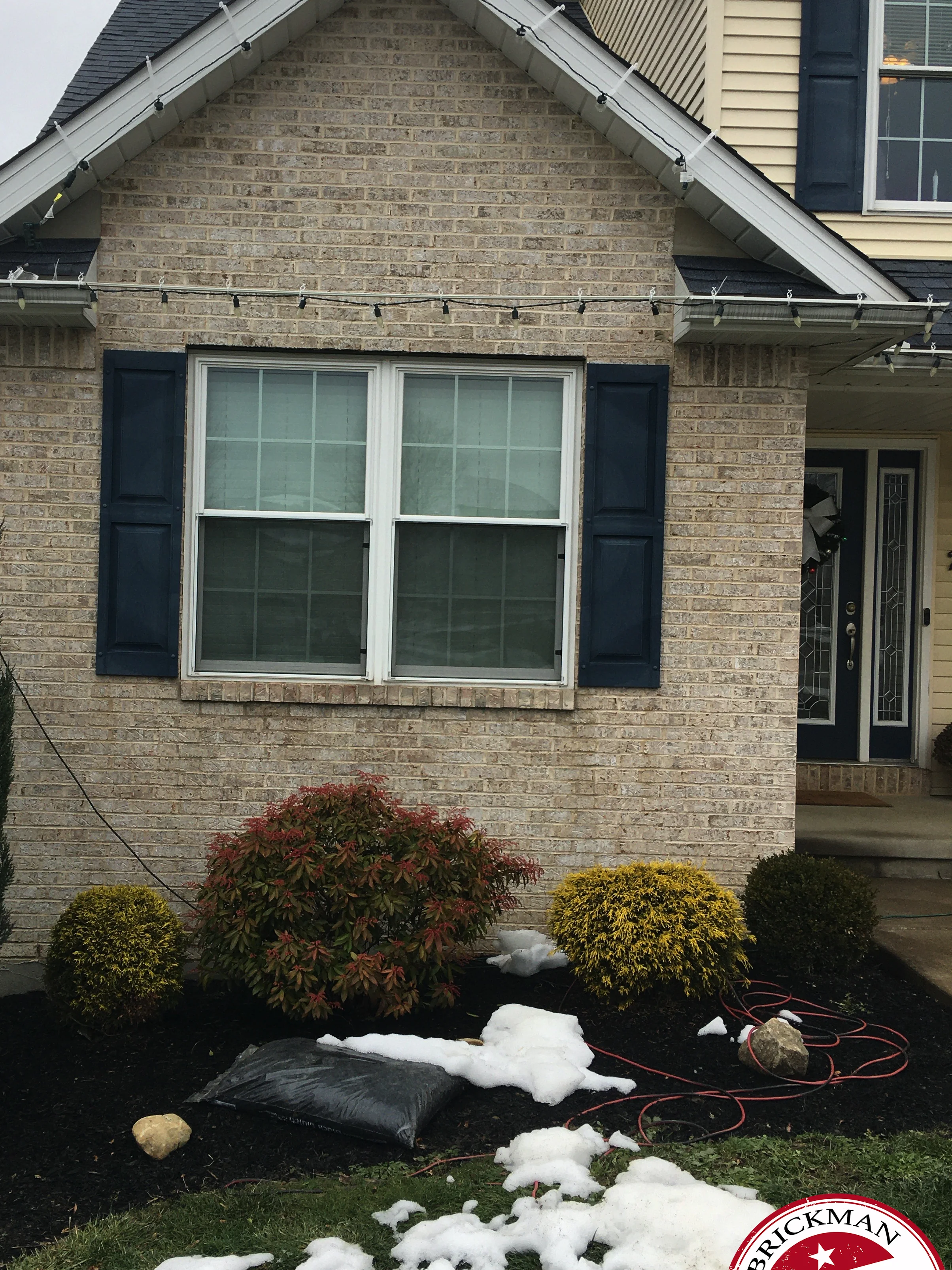 The gable end of the house in 2020 with snow on the lawn, one range of light brick from the peak to the course under the double window, and the certification stamp in the corner