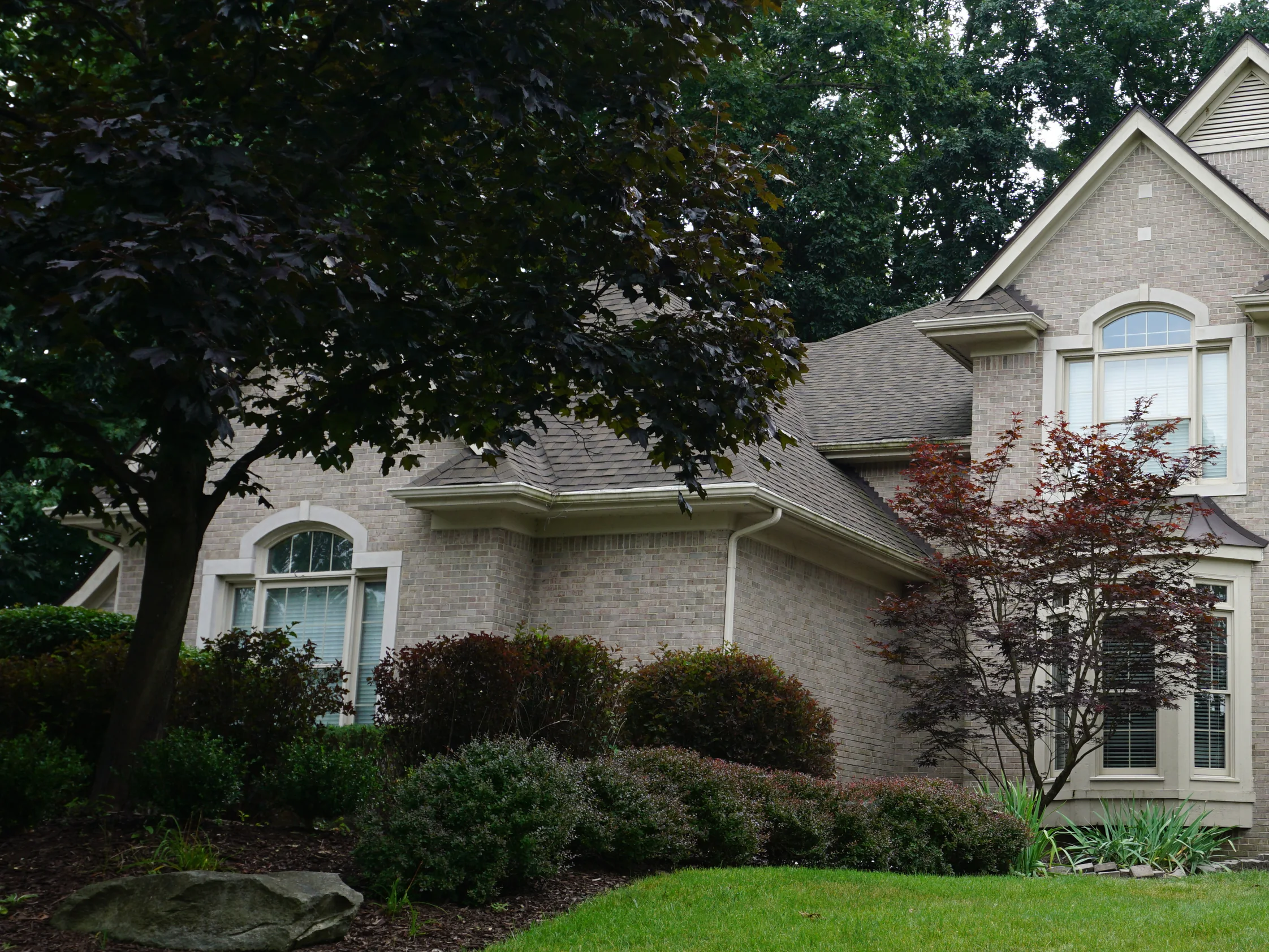 The house from the front lawn in 2019 under mature trees, the brick holding one buff and gray range across the wing, the gables and the bay