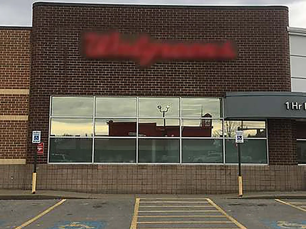 The whole storefront in 2021, dark red brown brick above a long band of shop window, a rounded brick corner at the right and marked accessible bays across the parking lot in front