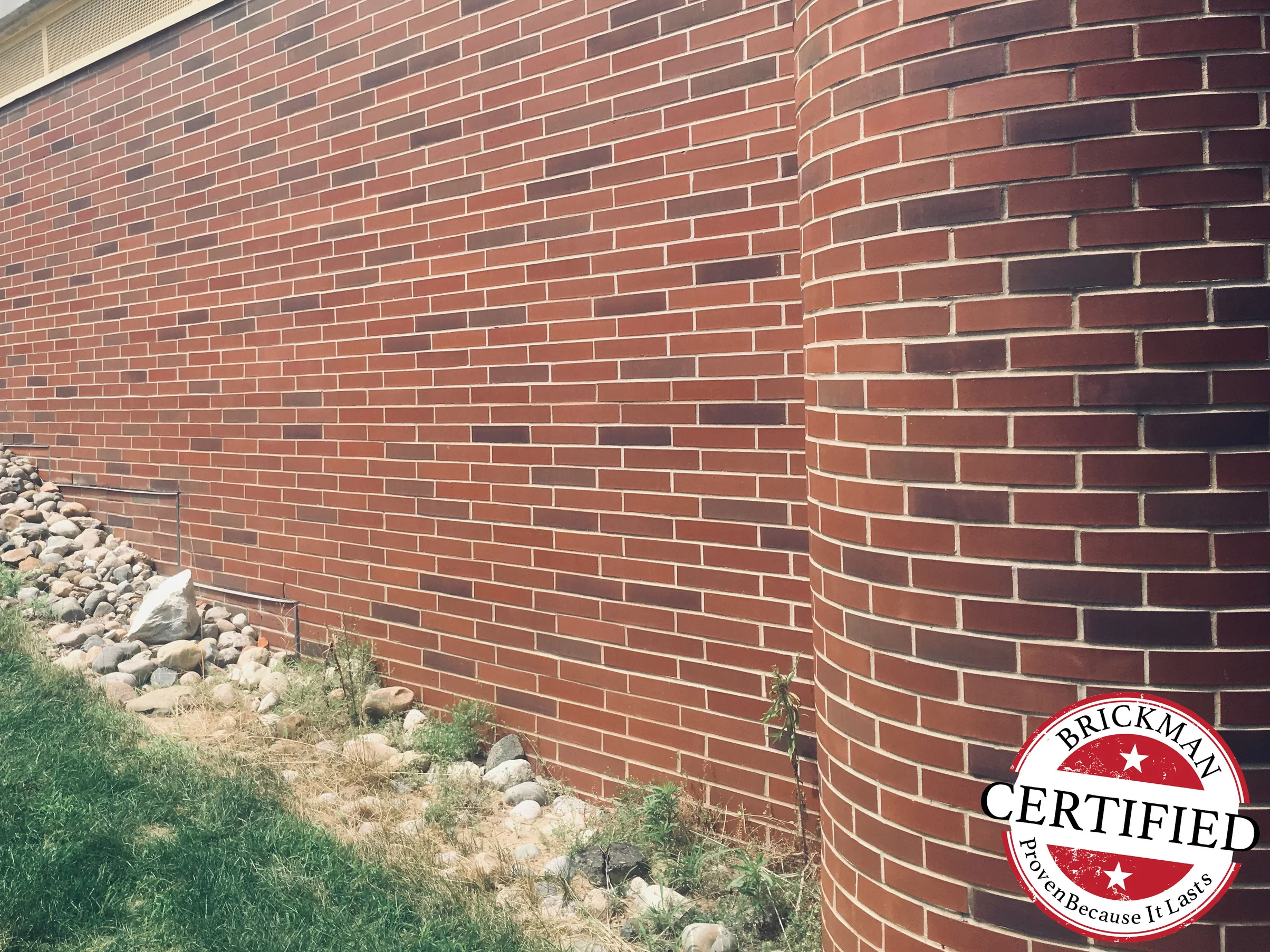 The same wall closer in 2019, running to a round brick column of radial units, maroon and dark flashed brick set through the red field, the frame carrying the Brickman Certified stamp