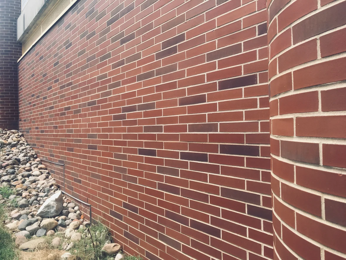 The stained wall in 2019 seen along its length to a round brick column, dark maroon units scattered through a red brick field above a stone bed