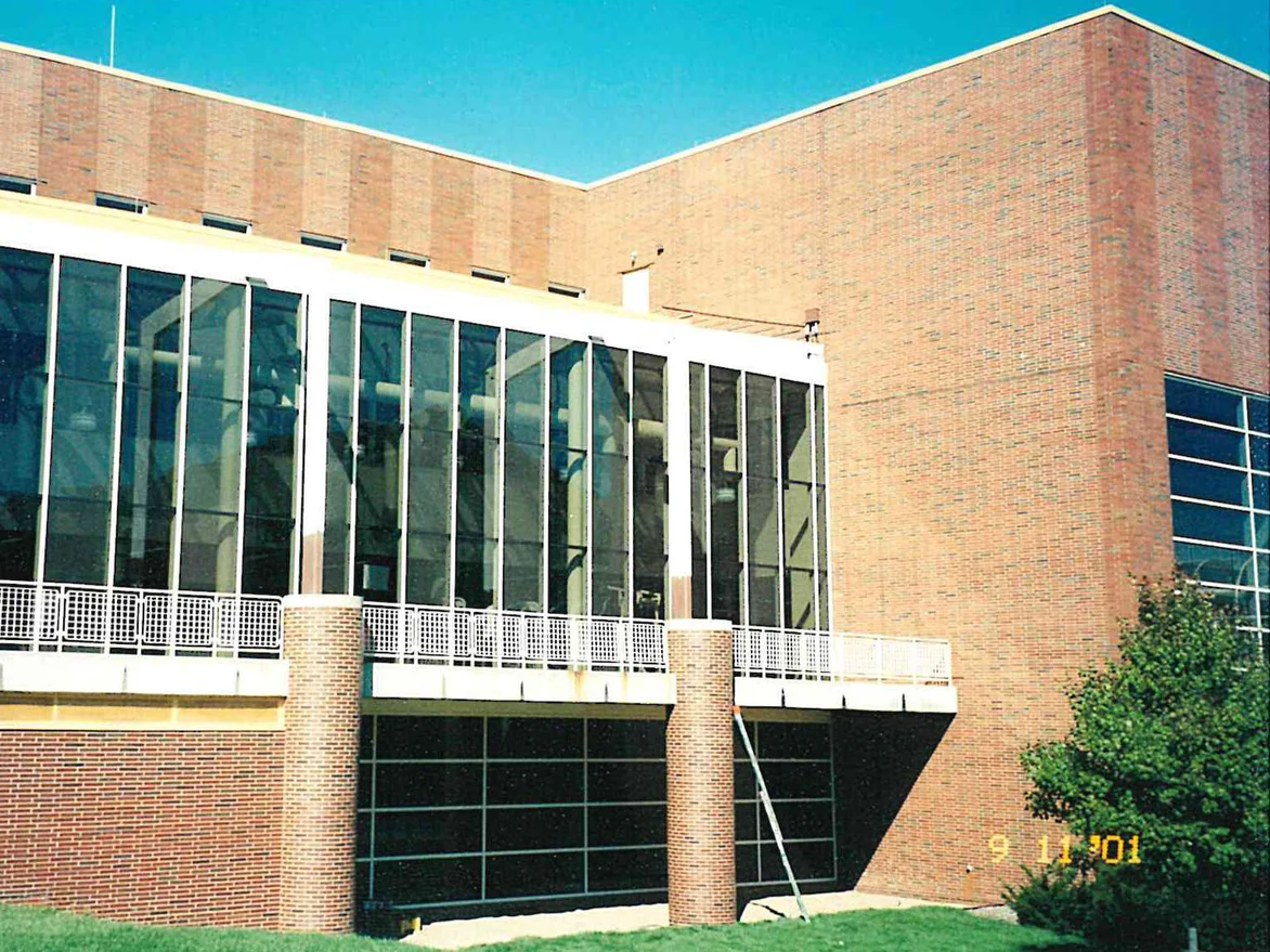 A glazed wing with a taller brick block behind it, a ladder standing against one of the round brick columns and a terrace rail above the brick base