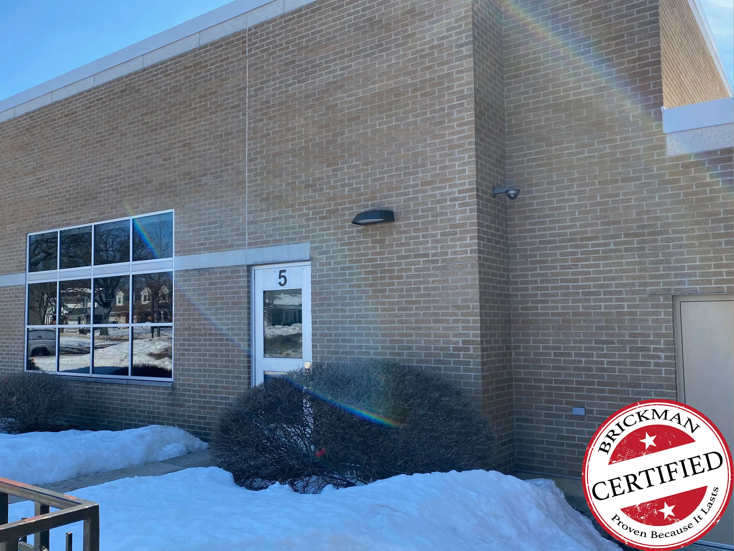An outside corner of the school in 2021 with snow on the ground, tan bark textured brick in one range around a door numbered 5, a wide mullioned window and a wall light, with the certification stamp in the corner