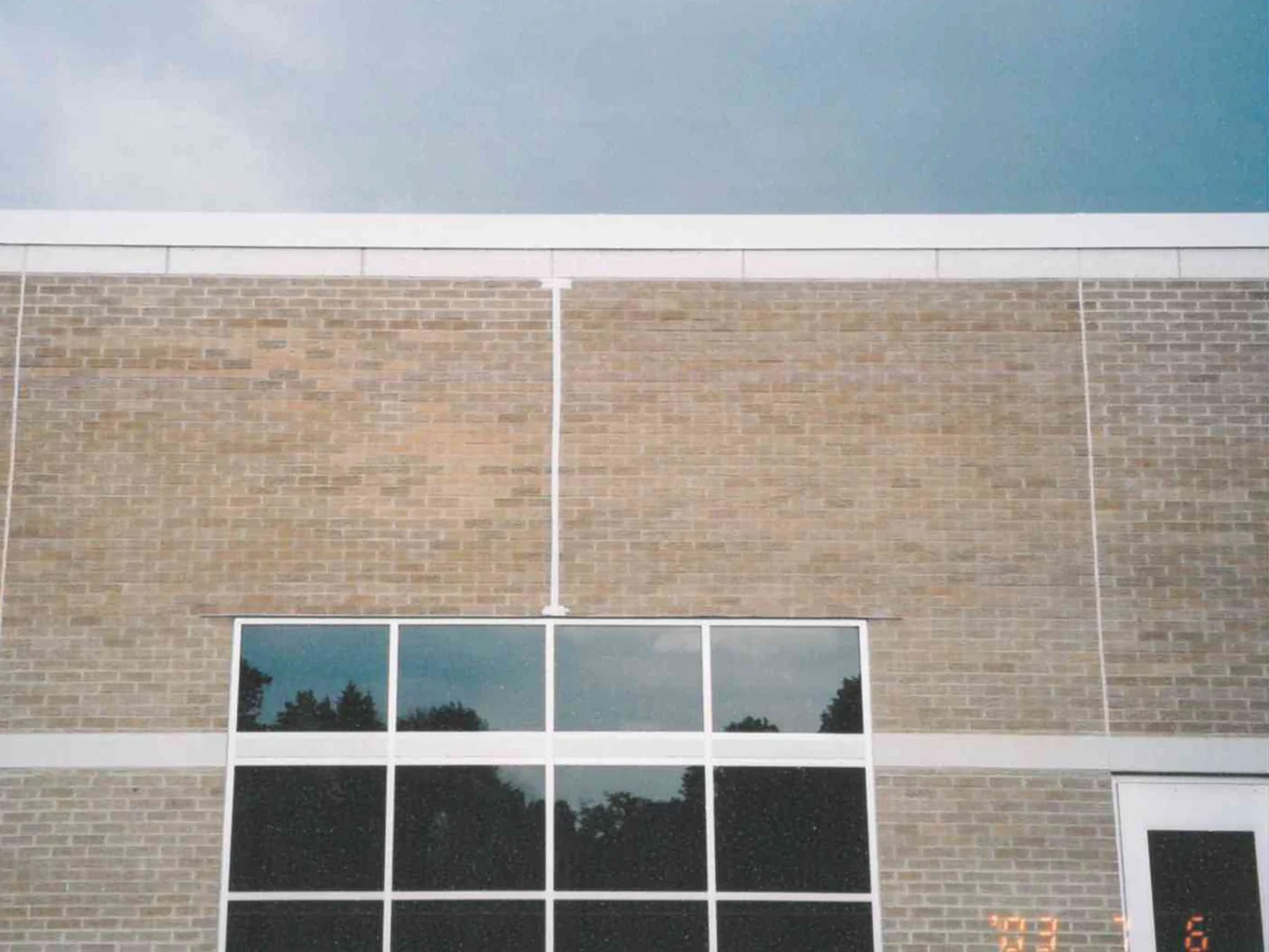 The upper wall of the school in July 2003 under a grey sky, tan bark textured brick with scattered darker units reading as blotches above a tall mullioned window, with a camera date in the corner