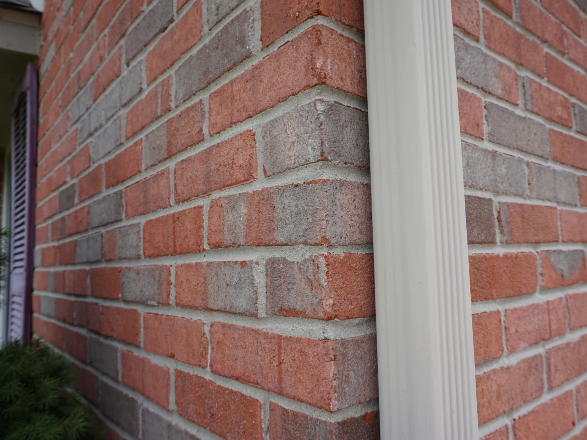 An outside brick corner of the house in 2019 with a white downspout running down it, the units in reds and greys with pale mortar joints
