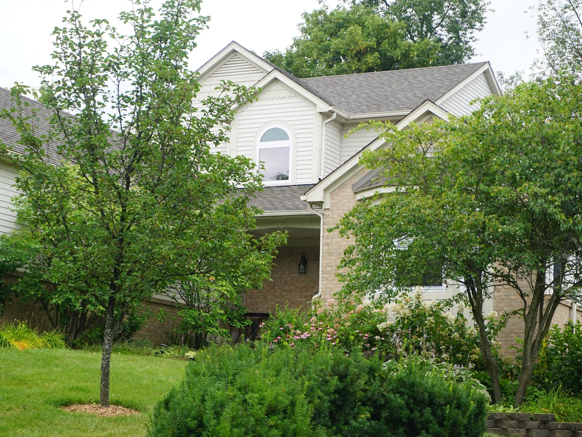 The front of the house in 2019 seen through two young trees, buff brick at the porch and the lower level under cream siding