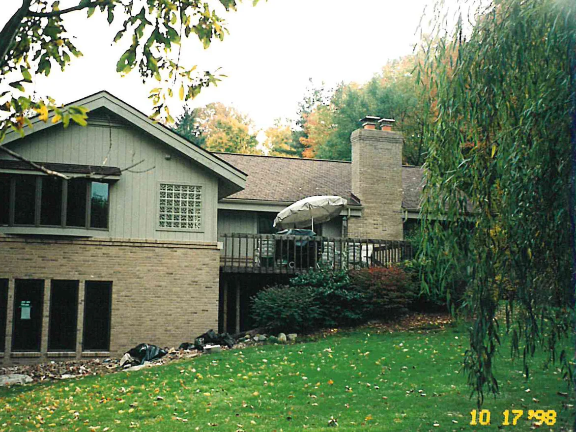 The rear of the house in 1998, a buff brick lower level under sage board and batten siding, with a raised deck and a willow at the right