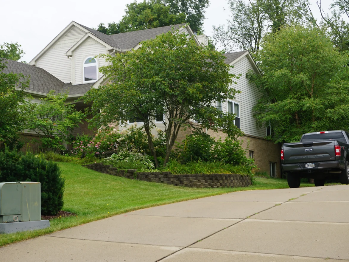 The house from the foot of the driveway in 2019, the buff brick of the walkout level running the length of the side under a bed of shrubs