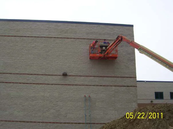 Two workers in an orange boom lift raised against the block wall in 2011, an extension ladder standing on the dirt below them