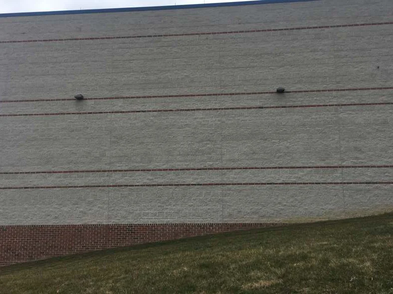 The block wall in 2021 under an overcast sky, one even pale range above a red brick base, with an established grass slope run up to the foot of it