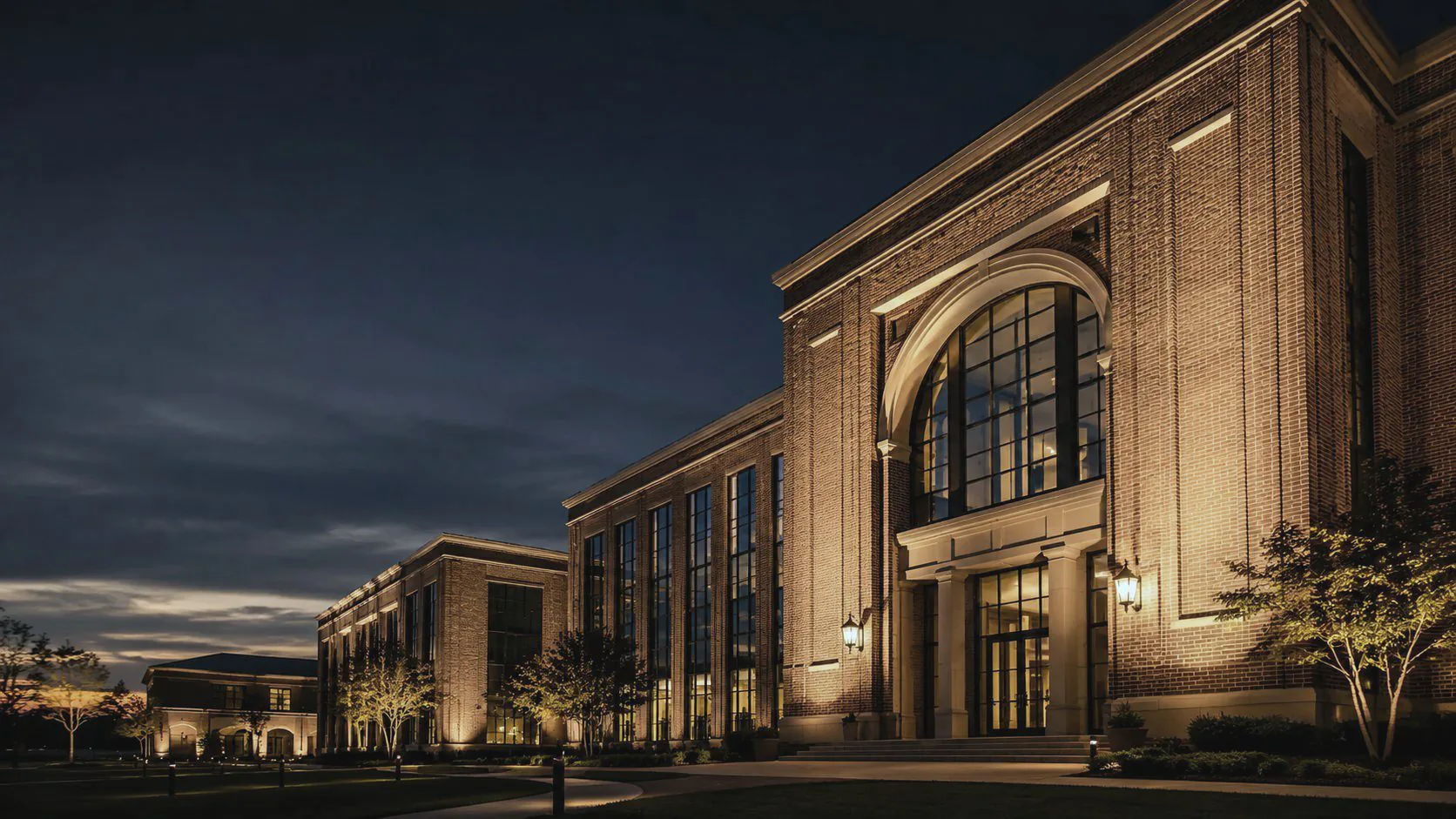 A long brick building at dusk, its arched entrance and tall windows lit from inside and below